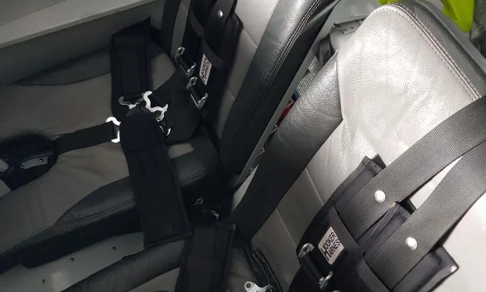 Two grey and black aircraft seats with multi-point harnesses.