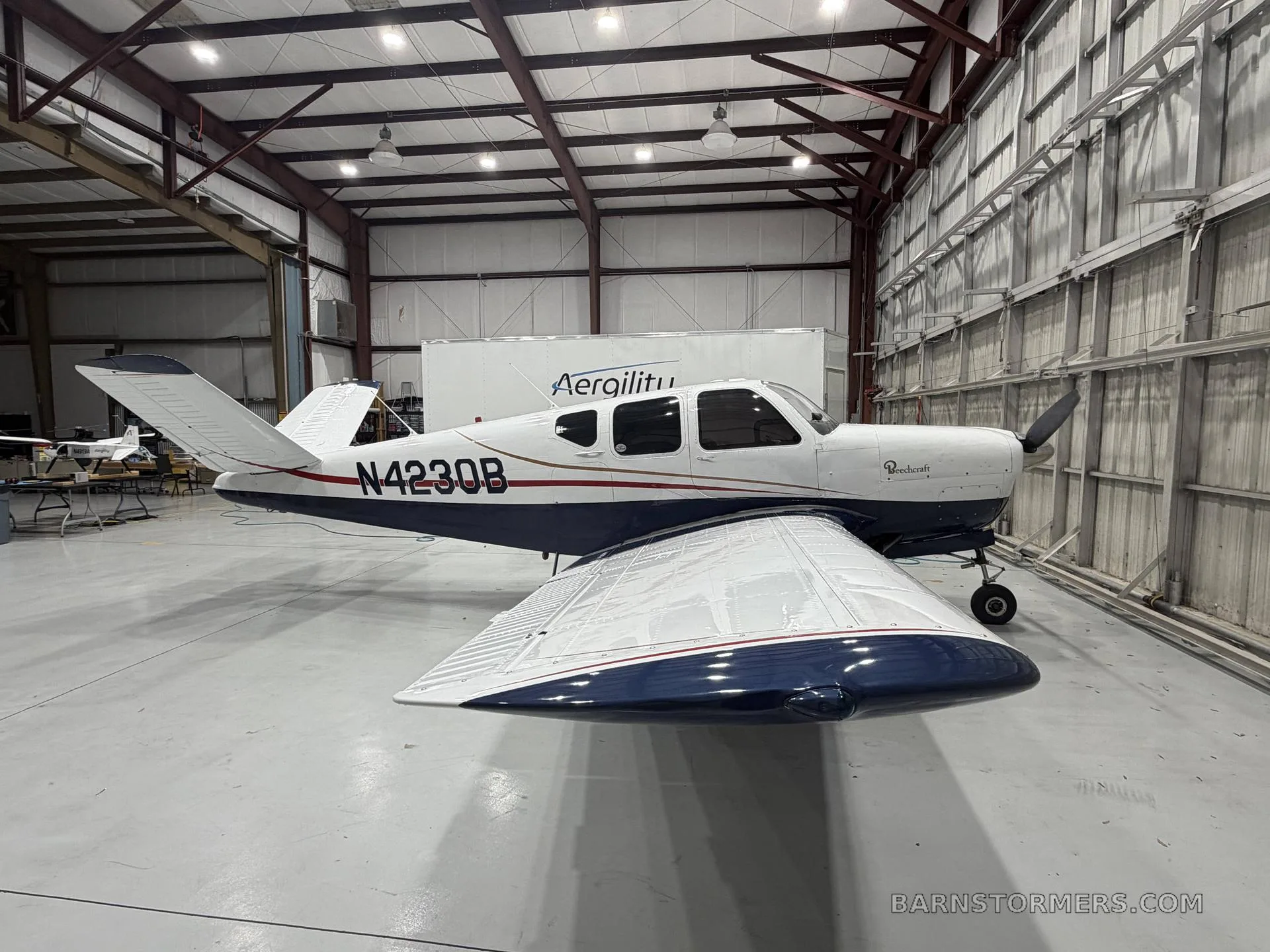 A white and blue Beechcraft Bonanza (N4230B) is parked inside a hangar, viewed from the front-left.