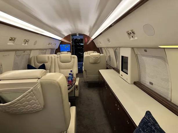 The luxurious interior cabin of a private jet features cream leather seats, dark wood cabinetry, and a galley area.