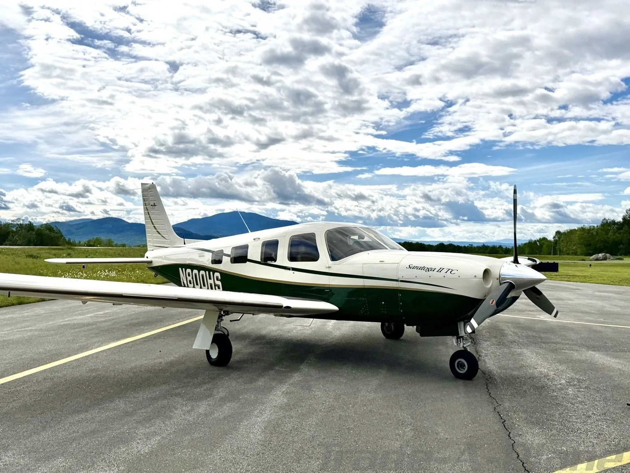 A white and green Piper Saratoga II TC aircraft, registration N800HS, parked on an asphalt ramp under a bright, cloudy sky.