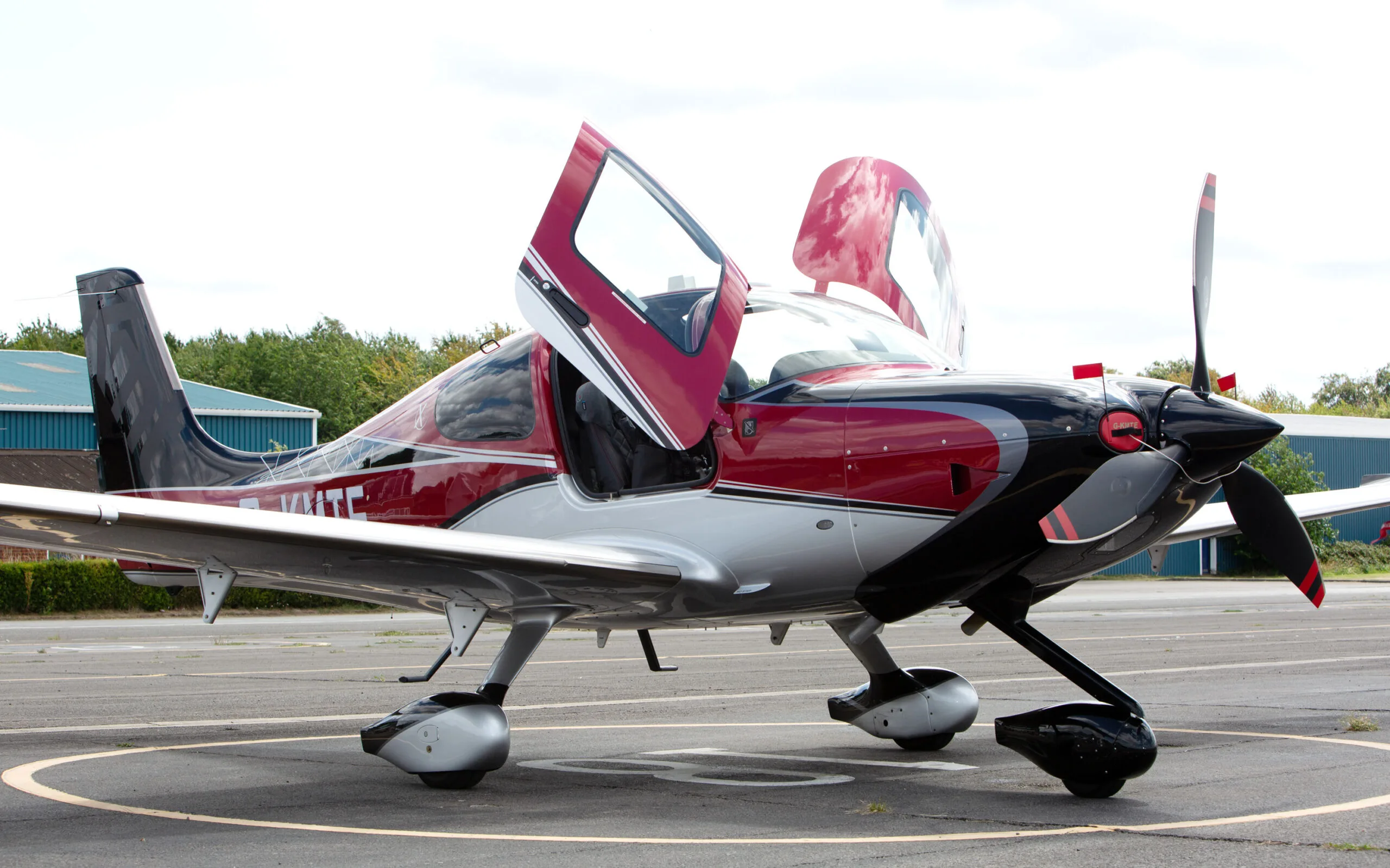 A three-quarter front view of a red, black, and silver Cirrus SR22 aircraft with its gull-wing doors open, parked on an airfield.