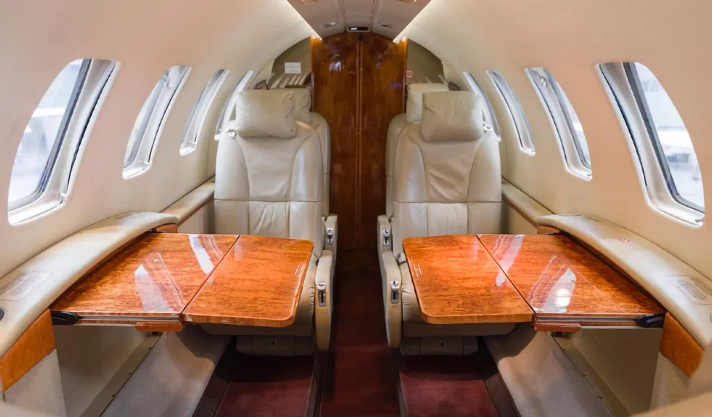 The aircraft cabin features two rows of beige leather seats facing each other with polished wooden fold-out tables.