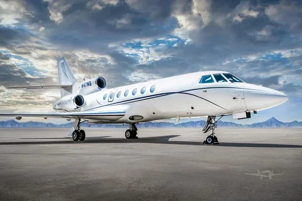 A white Dassault Falcon 900EX business jet with blue stripes, registration N47NS, on a tarmac with a cloudy sky and mountains in the background.