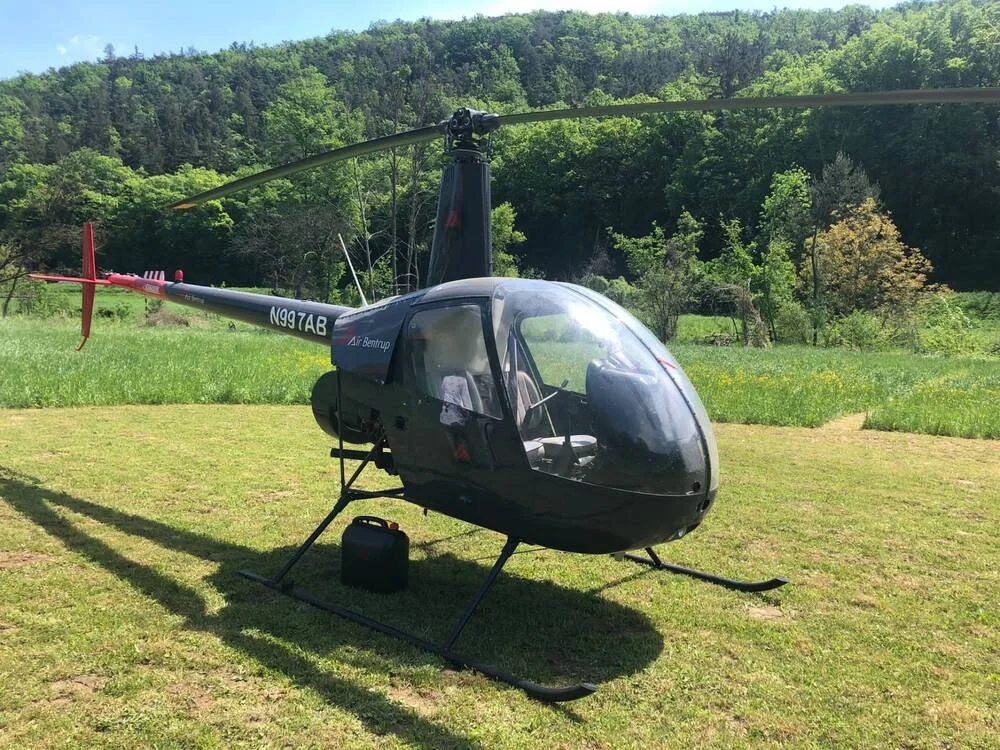 A dark gray Robinson R44 helicopter, N997AB, with red tail accents, parked in a green grassy field with trees in the background.