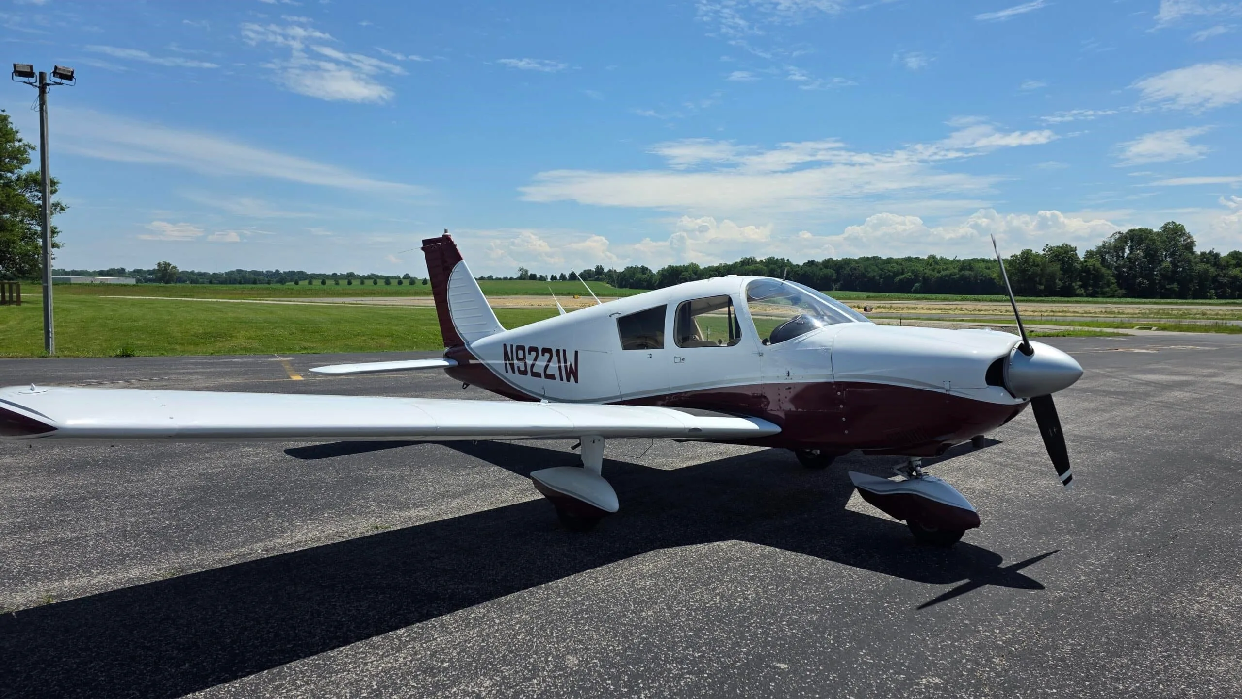 A white and maroon Piper Cherokee aircraft (N9221W) parked on an asphalt ramp under a blue sky.
