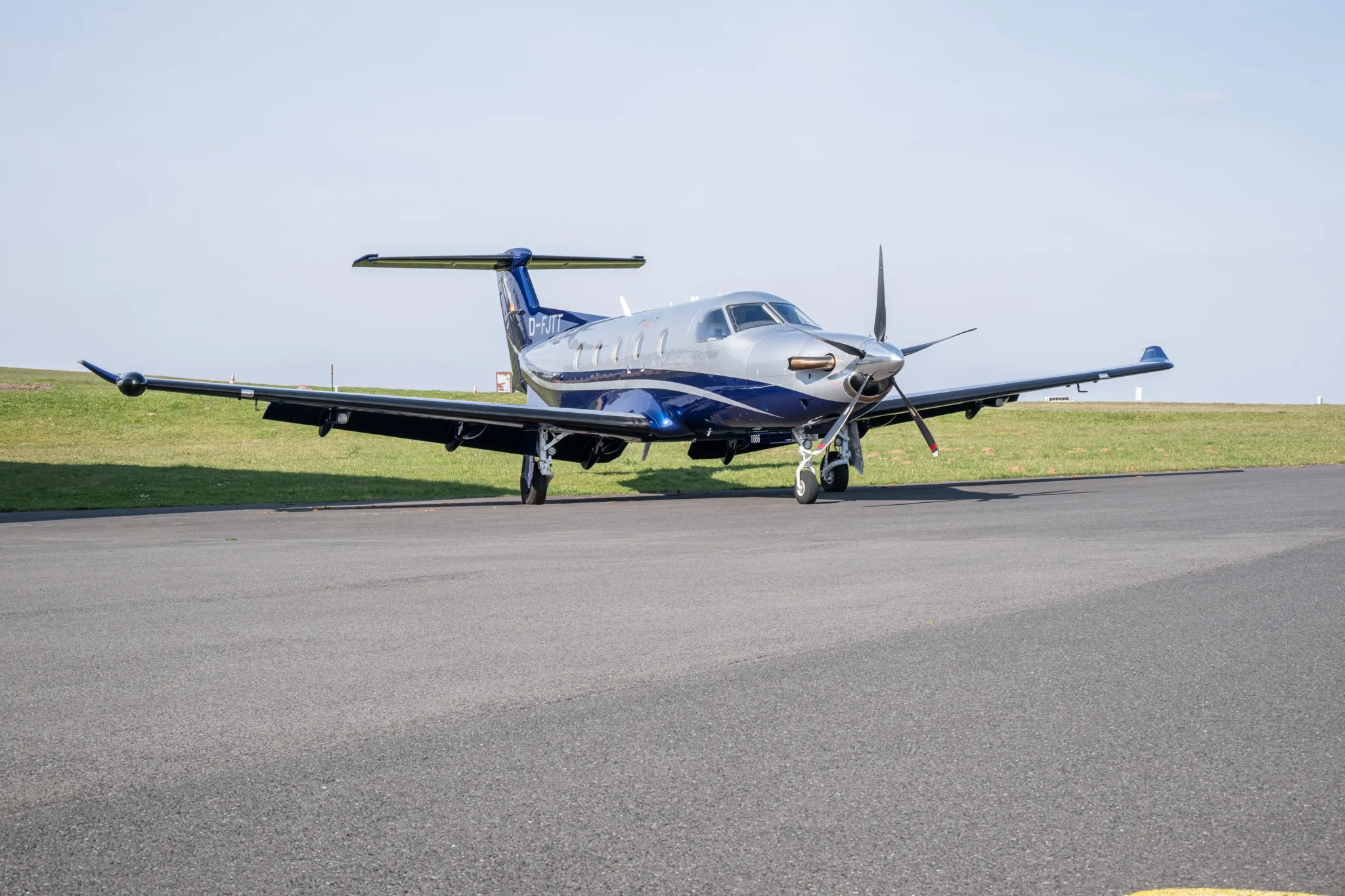 A silver and blue Pilatus PC-12 NG aircraft is parked on an asphalt runway under a clear sky.