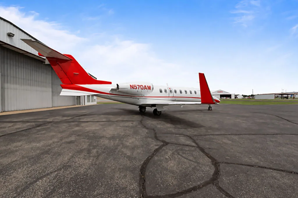 A white and red business jet (N570AM) seen from the rear, parked on an asphalt tarmac next to a hangar.