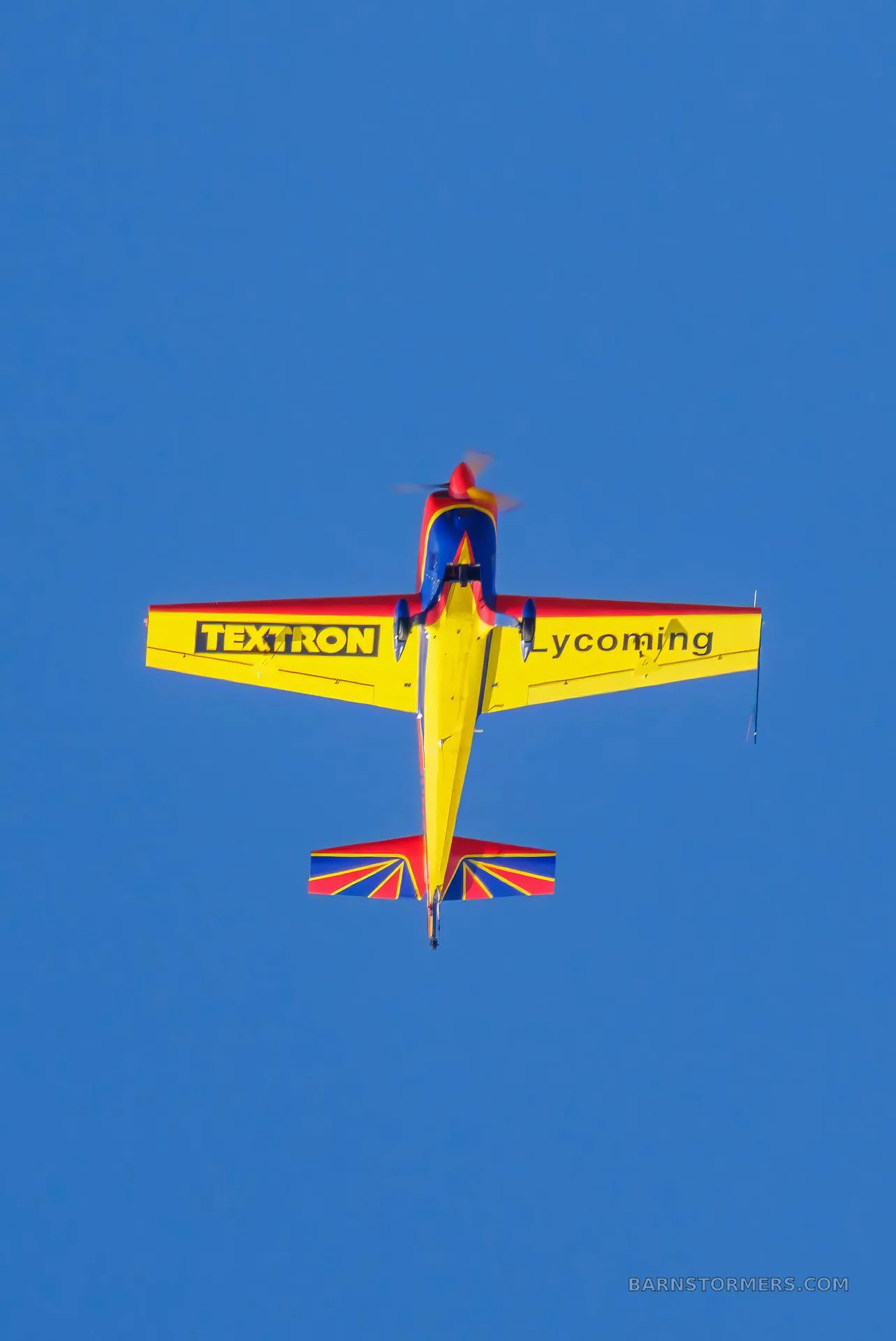 A red, blue, and yellow aerobatic aircraft is seen flying inverted from a top-down view against a clear blue sky, showing "TEXTRON" and "Lycoming" on its wings.