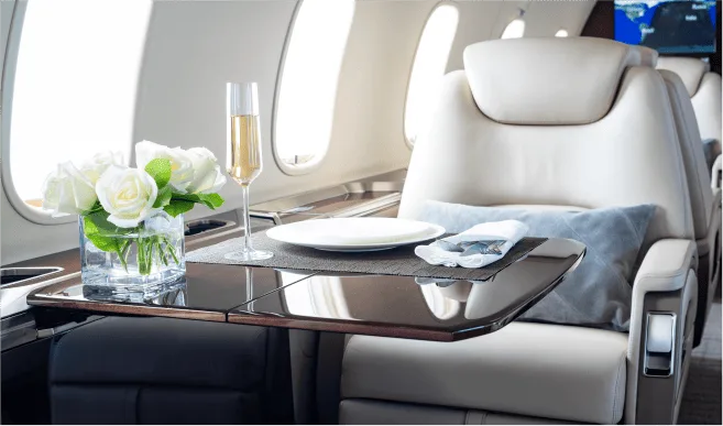 A private jet cabin table is set with a plate, silverware, a glass of champagne, and a vase of white roses.