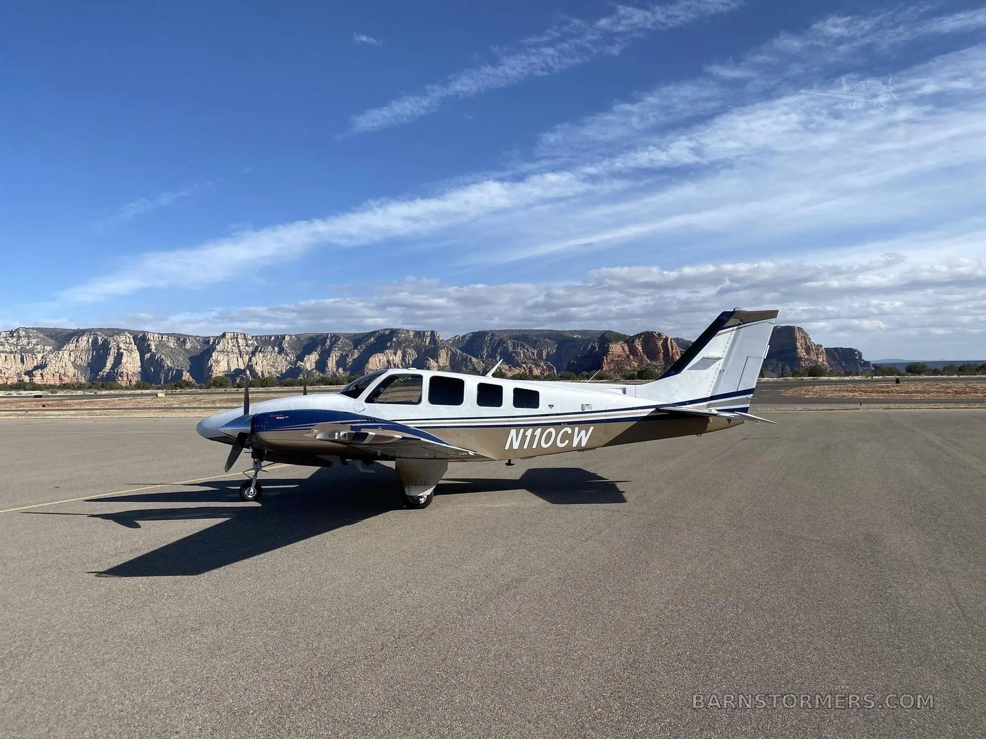 A white and blue twin-engine aircraft with tail number N110CW is parked on an asphalt runway with red rock mountains in the background under a partly cloudy sky.