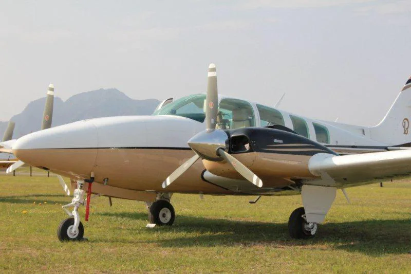 A close-up front view of a white and gold twin-engine propeller aircraft parked on a grassy field with mountains in the background.