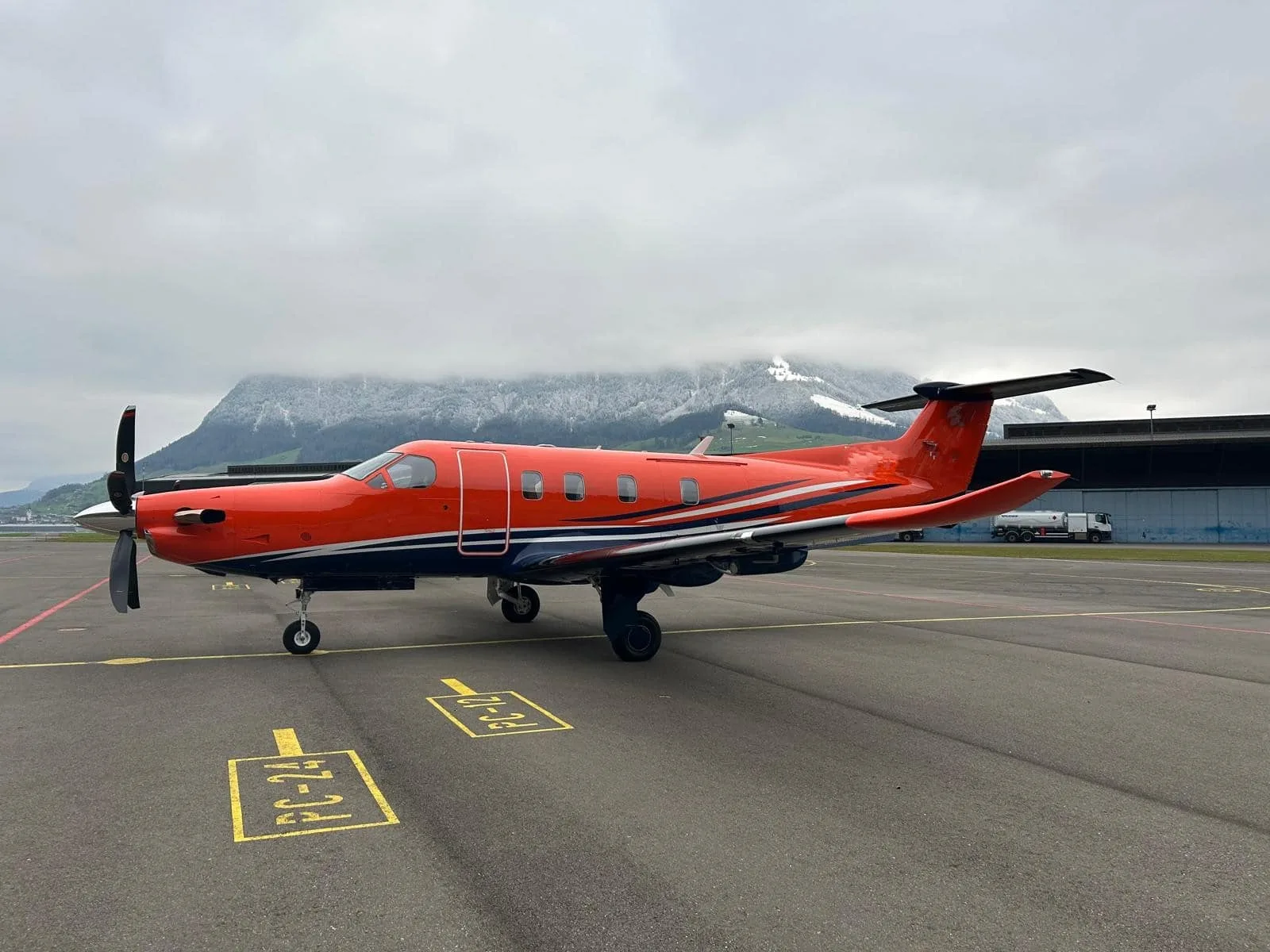 A bright orange Pilatus PC-12 aircraft with dark blue and white stripes sits on an airport tarmac with snow-capped mountains in the background.