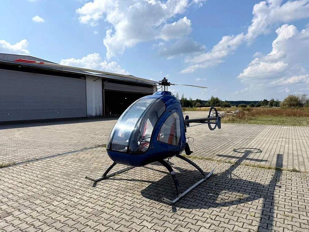 A blue single-seat helicopter with a clear bubble canopy sits on a paved area outside a hangar under a partly cloudy sky.