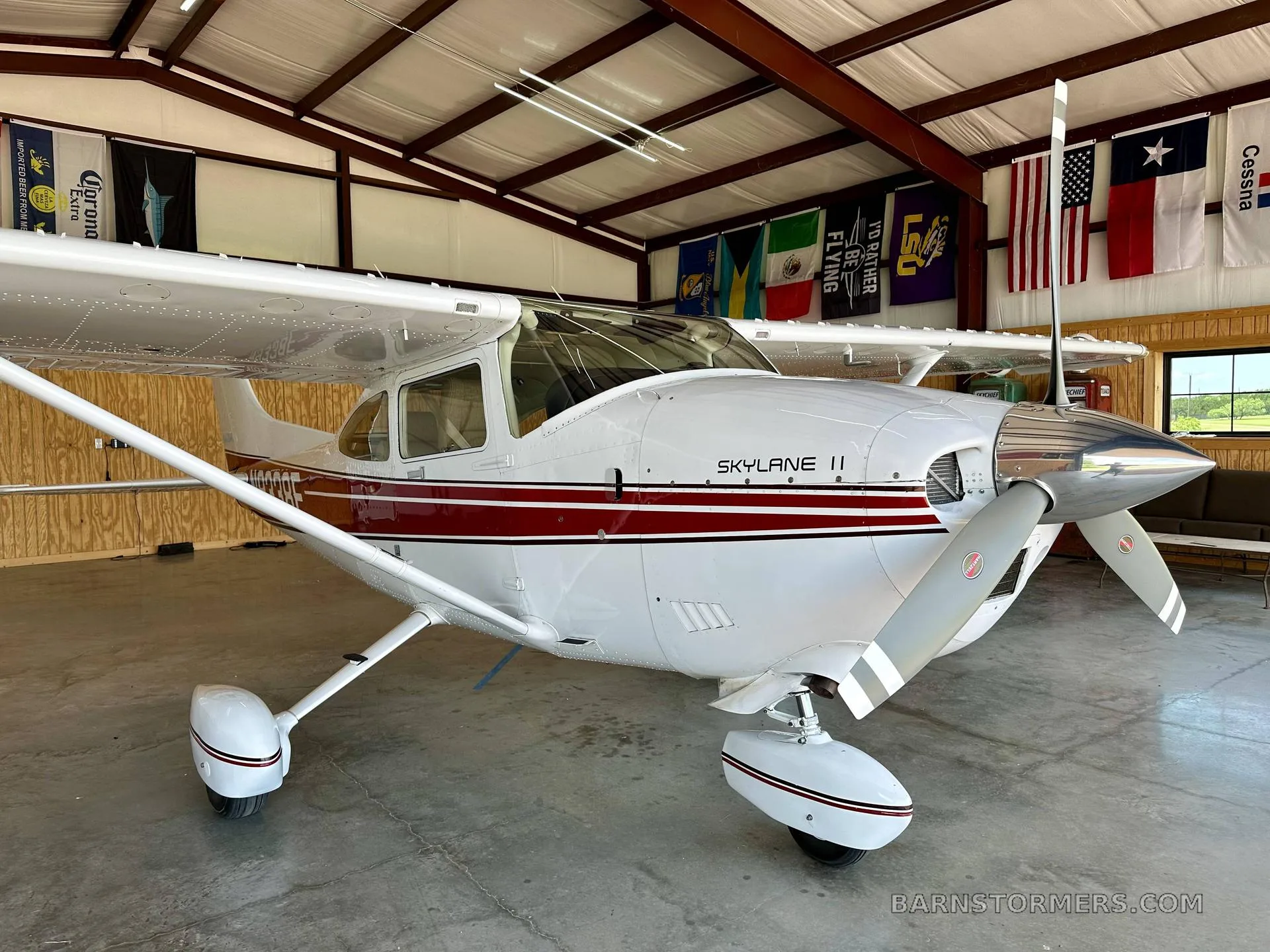 The front-right view of a white Cessna Skylane II with red stripes and a polished propeller spinner, parked in a hangar.