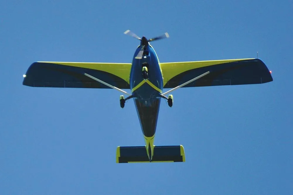 A yellow and blue Skyper GT9 aircraft flying against a clear blue sky, viewed from below.