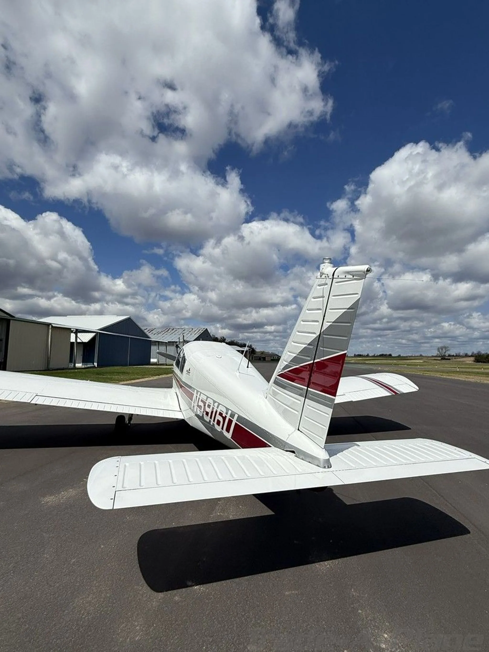 A rear view of a white Piper Cherokee aircraft with red and grey stripes parked on an airfield.