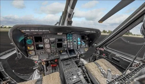 The cockpit of a helicopter features multiple screens, gauges, and controls, with a view of the tarmac outside.