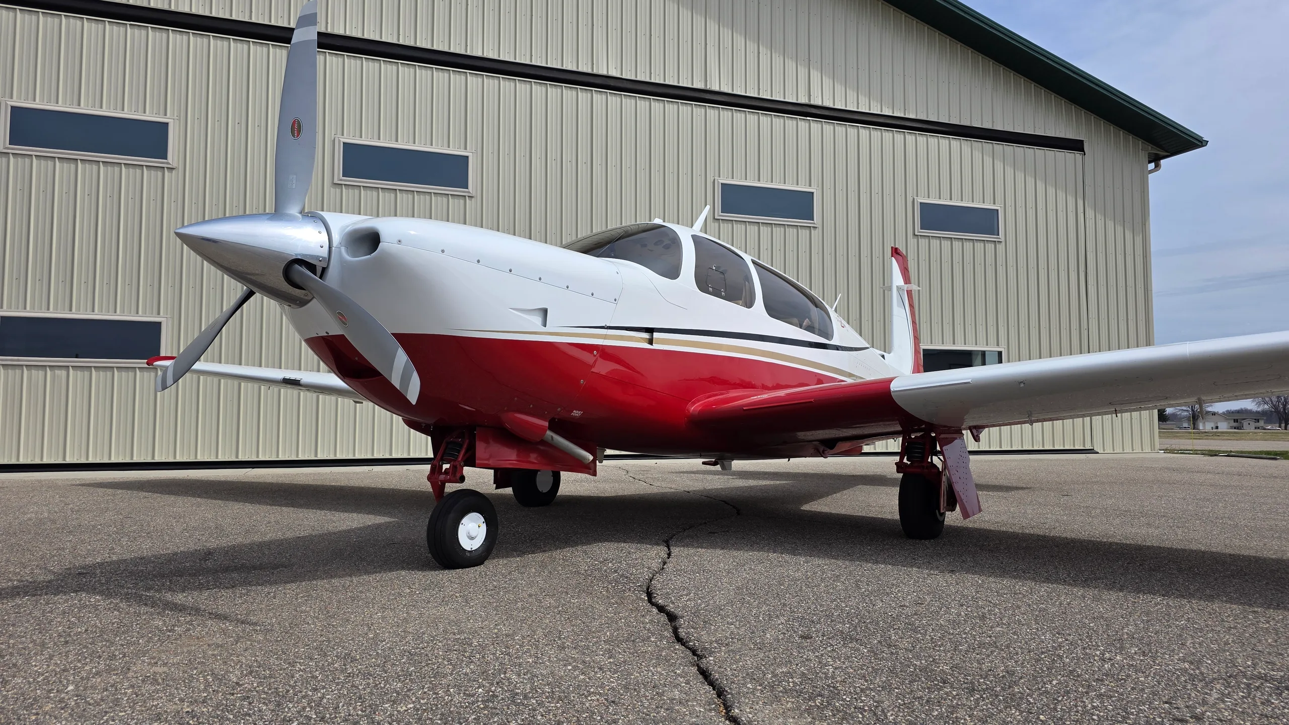 A red and white Mooney aircraft with gold stripes parked on an asphalt apron next to a hangar.