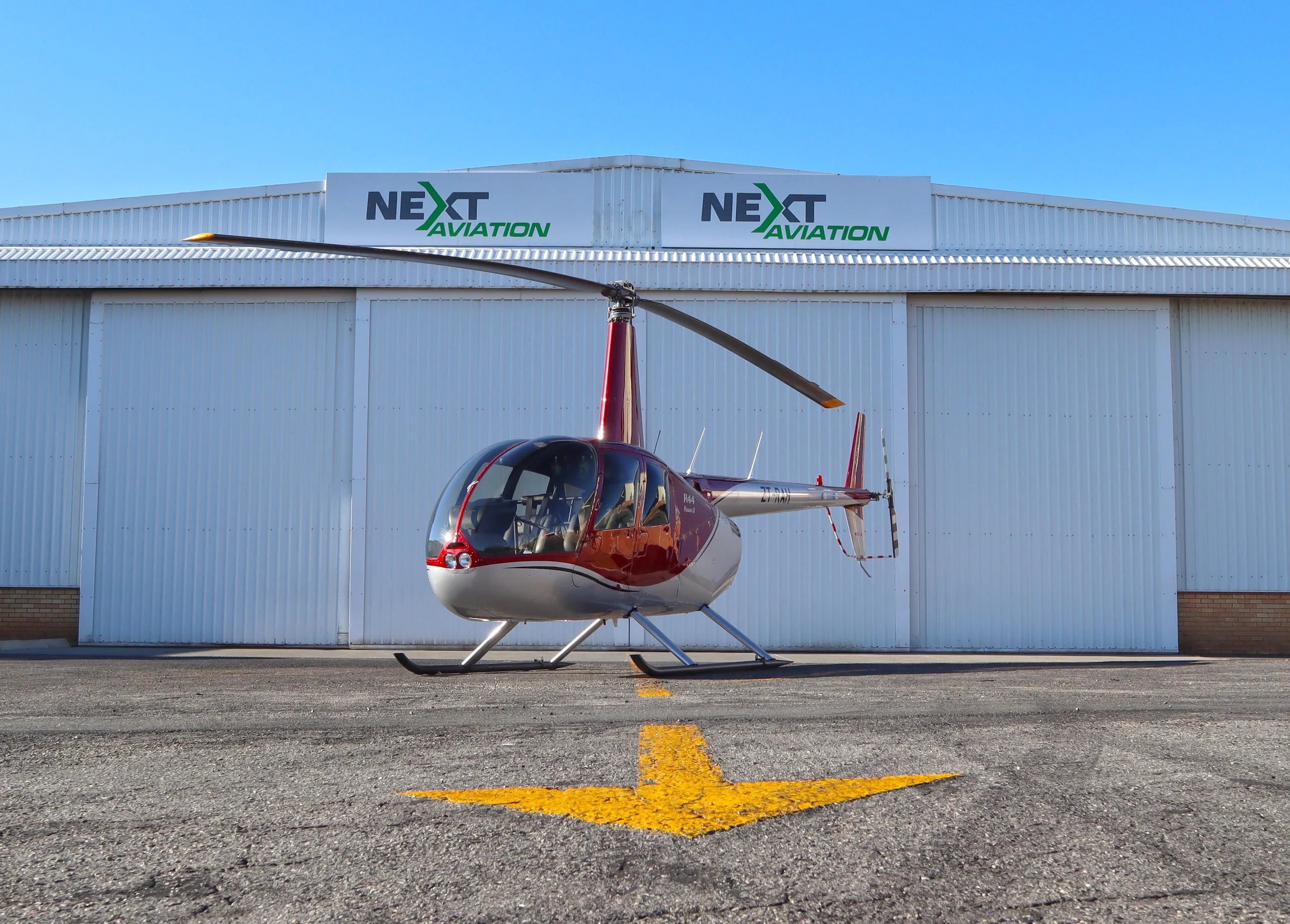 A red and silver Robinson R44 Raven II helicopter is parked on an asphalt tarmac in front of a white hangar with 'NEXT AVIATION' logos.