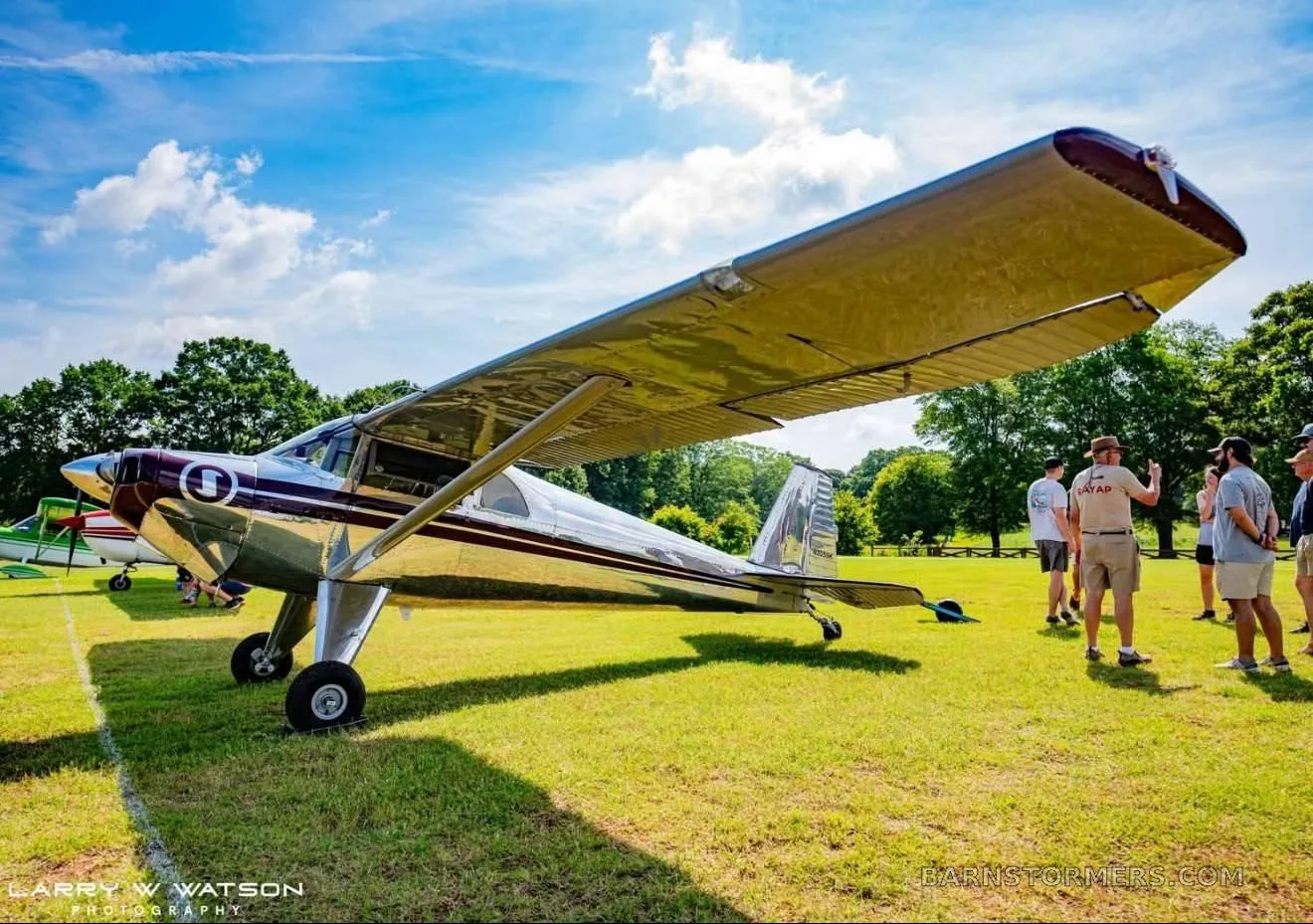 A polished silver high-wing aircraft with maroon accents on the nose and wingtips, parked on a grassy field with people nearby.