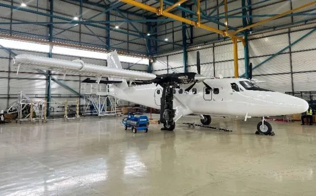 A white twin-engine turboprop aircraft is shown from the side in a hangar, with its engine cowling open and a service cart nearby.