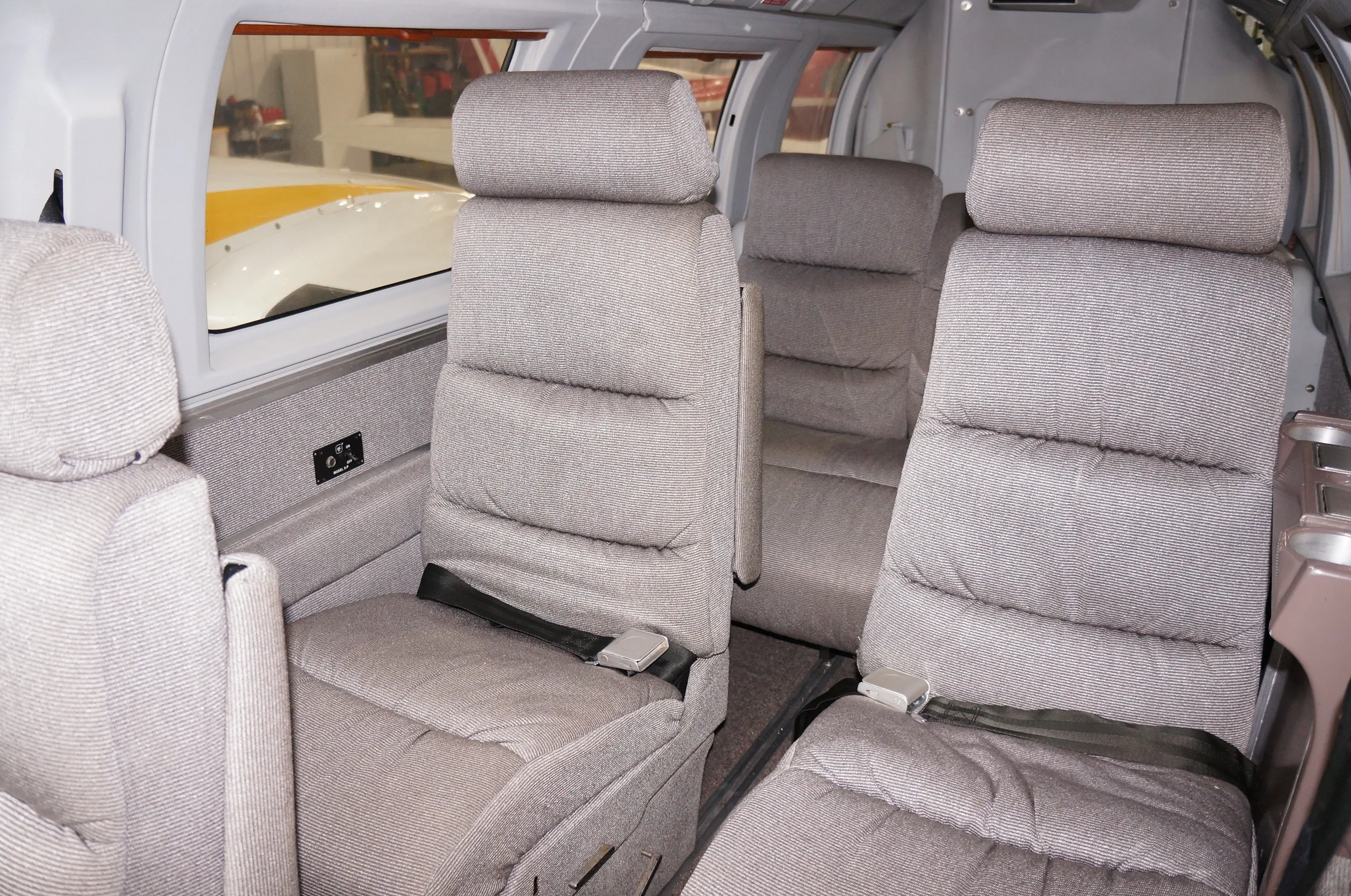 The aircraft cabin features two rows of light gray fabric seats with seatbelts, looking towards the front.