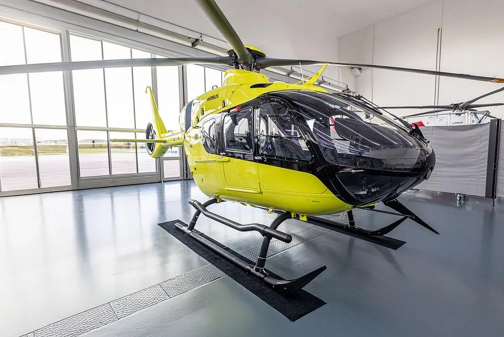 A bright yellow Airbus H135 helicopter with black accents is parked inside a hangar, viewed from the front-left.