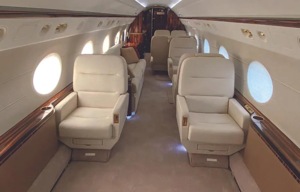 The luxurious interior cabin of a private jet, featuring cream-colored individual seats and a sofa, with polished wood accents.