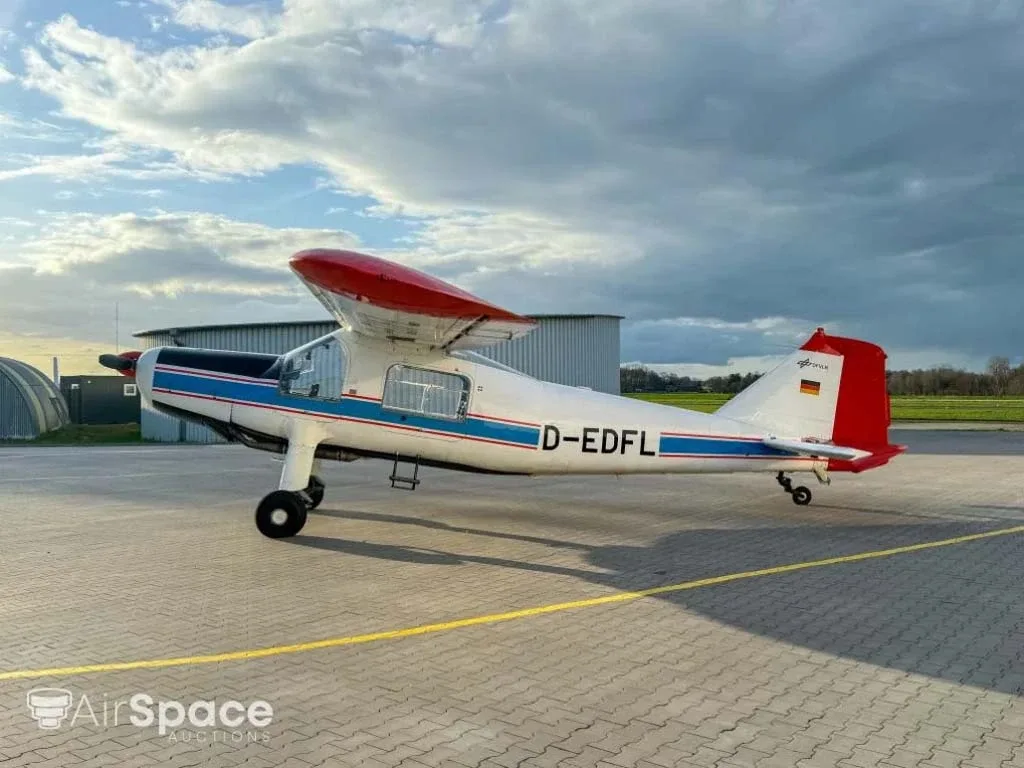 A side view of a white aircraft with blue and red stripes and red wingtips, parked on a paved apron under a cloudy sky.