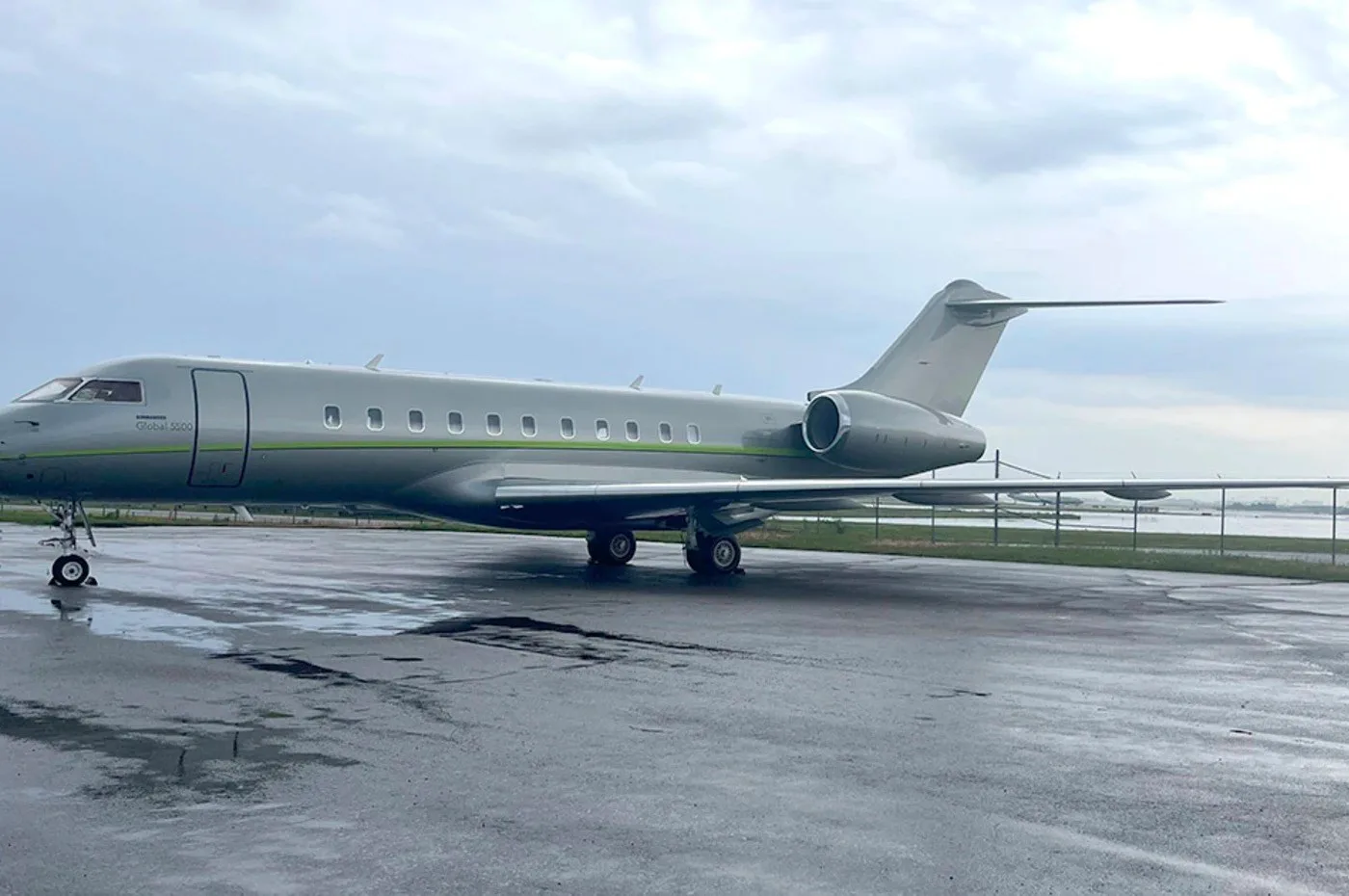 A grey Bombardier Global 5000 business jet with a green stripe on a wet tarmac under a cloudy sky.