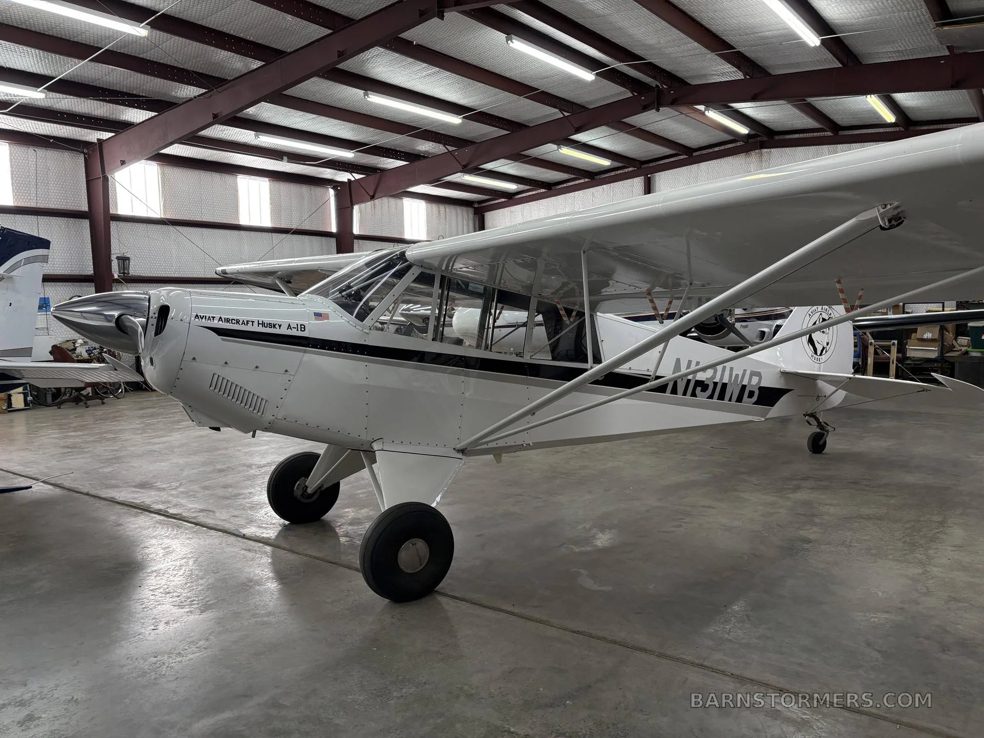 A three-quarter front view of a white Aviat Aircraft Husky A-1B with black trim and tail number N131WB, parked in a hangar.