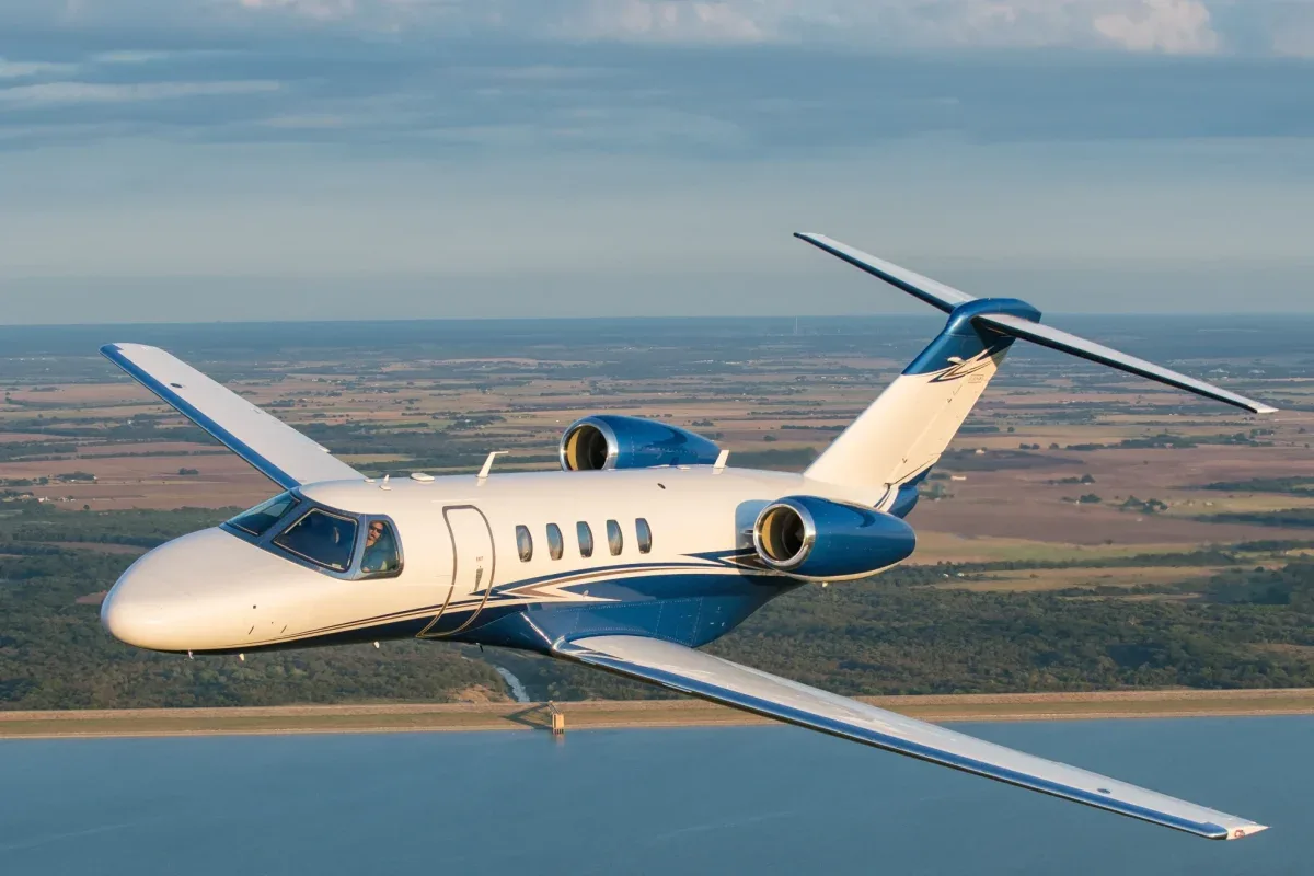 A blue and white Cessna Citation CJ4 flying over a lake.