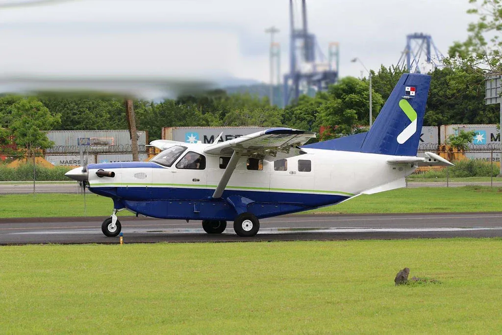 A blue and white Quest Kodiak 100 aircraft with a green tail logo on a runway, with industrial cranes in the background.