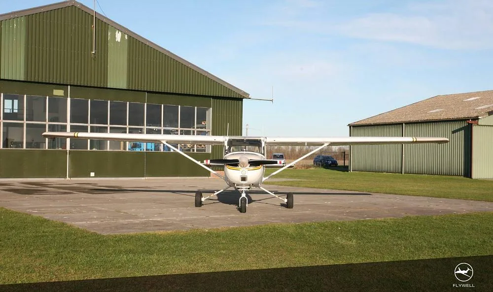 A head-on view of a white and beige Cessna 172 parked on a concrete pad in front of a large green hangar.