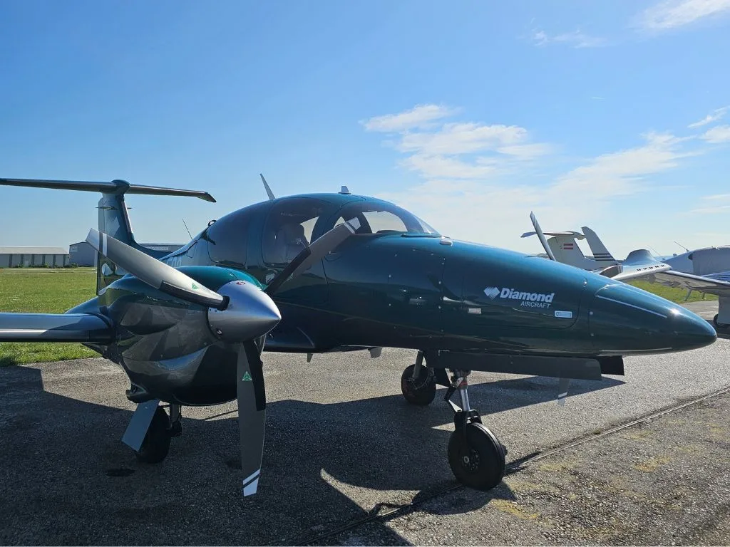 A dark green Diamond DA62 aircraft is parked on a tarmac, viewed from the front-right, with other aircraft in the background.