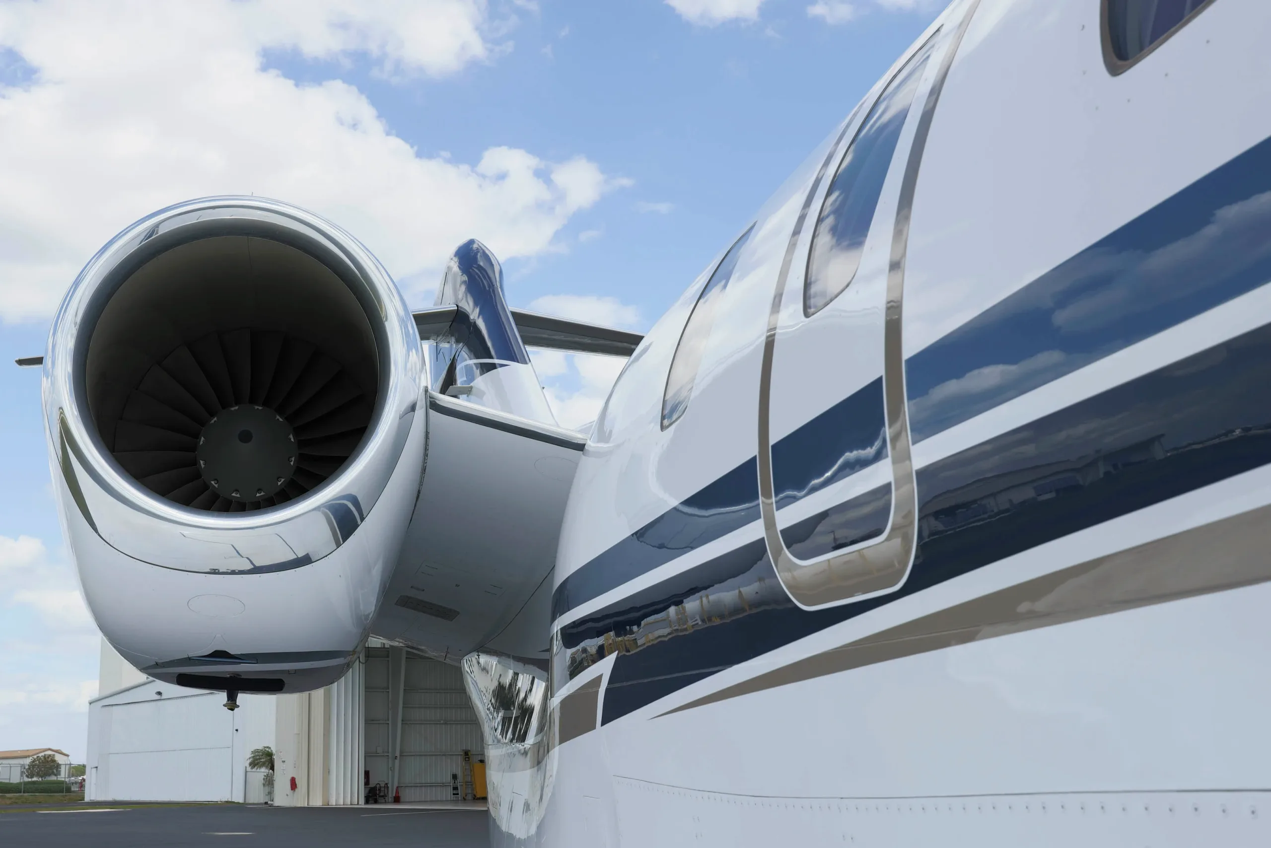 A close-up view of the engine and fuselage of a white business jet with blue and grey stripes, parked near a hangar.