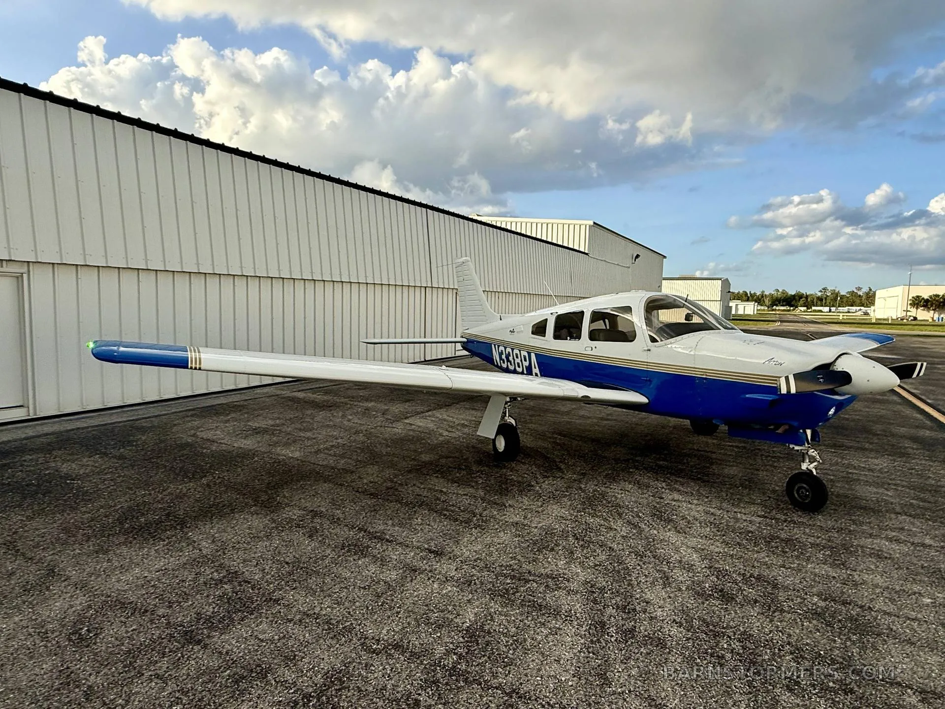 A white and blue Piper Cherokee aircraft (N338PA) is parked on an asphalt ramp with a hangar in the background under a cloudy sky.