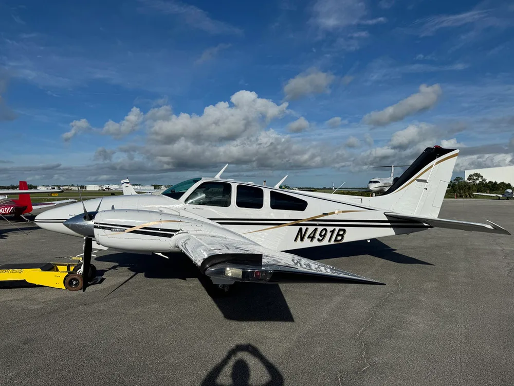 A white twin-engine Beechcraft Baron aircraft with black and gold stripes, registration N491B, parked on an airfield under a partly cloudy sky.