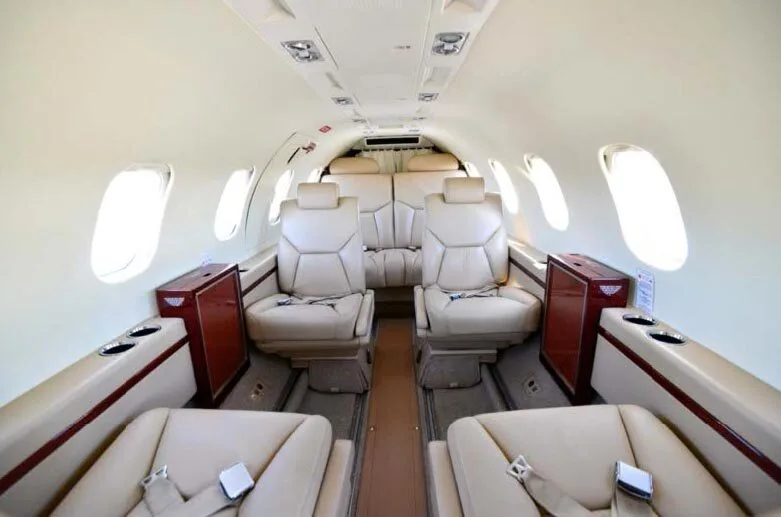 An aircraft cabin interior featuring light tan leather seats and dark wood trim, viewed looking towards the rear.
