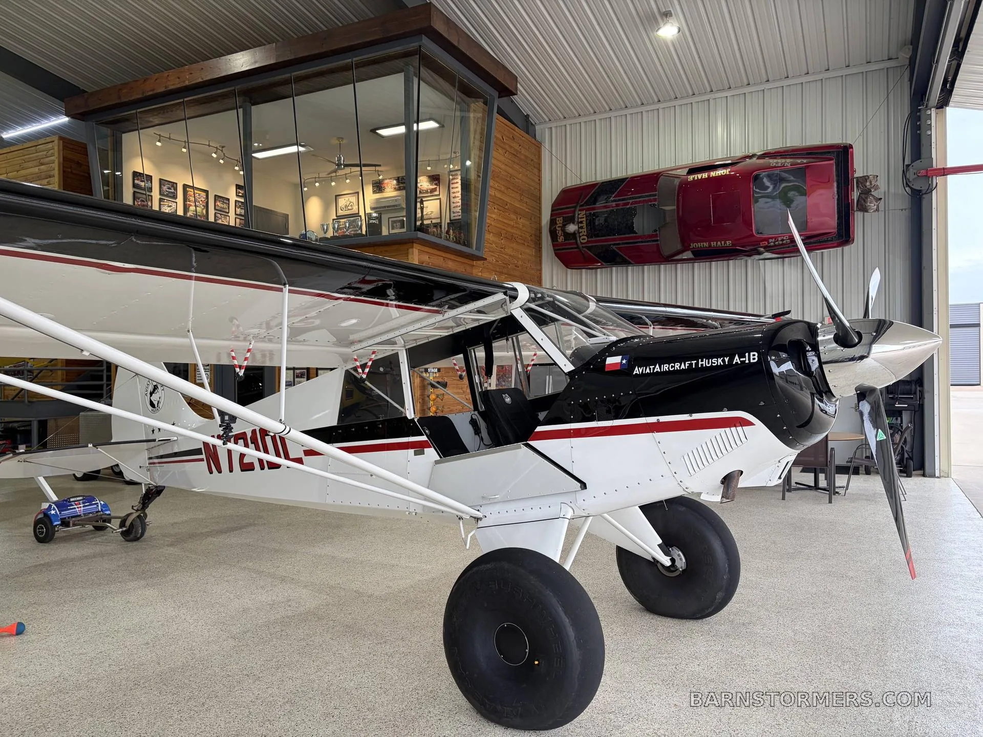 A black and white Aviat Aircraft Husky A-1B with red stripes and large tundra tires, parked in a hangar with a red classic car mounted on the wall.