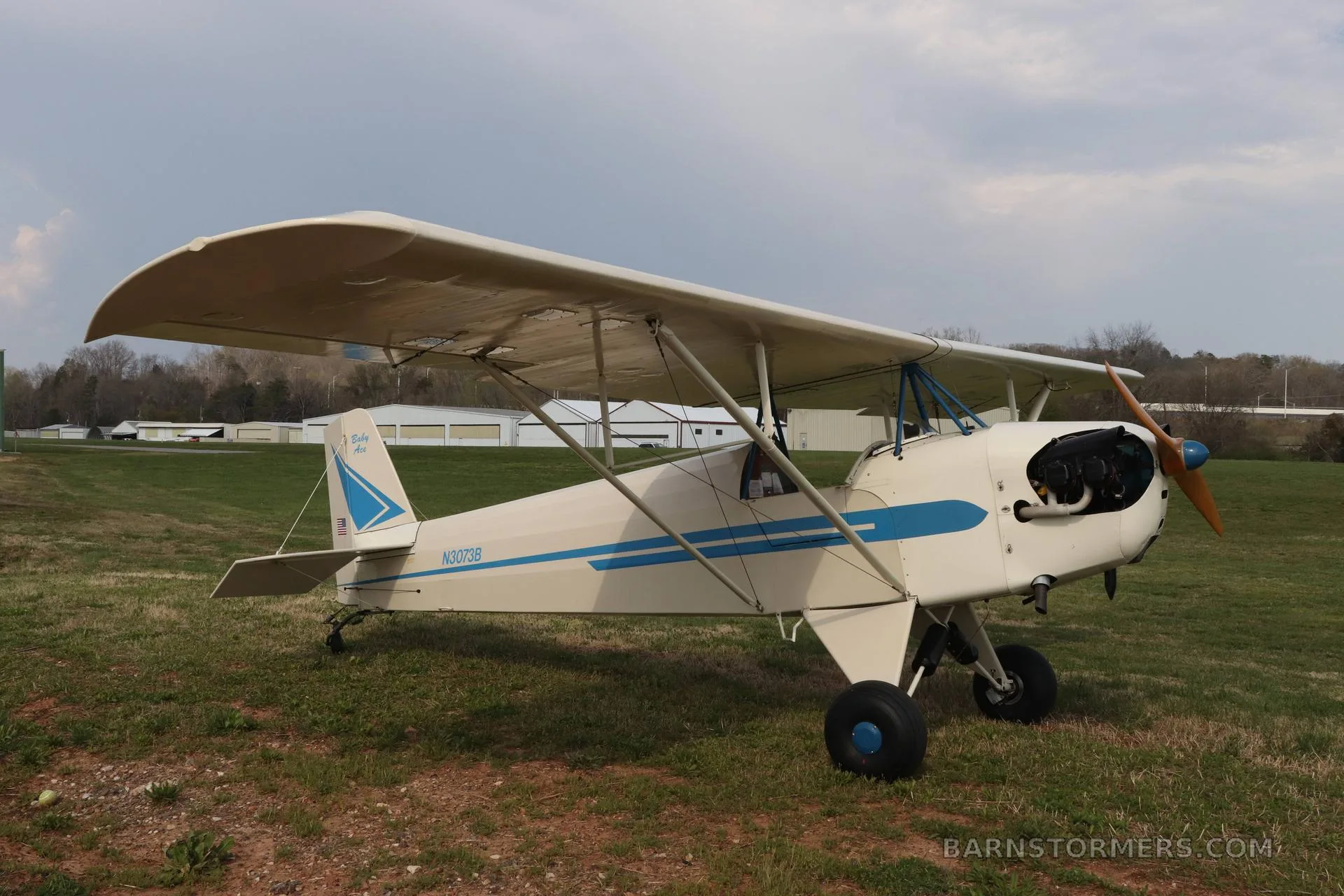 A white and blue single-engine taildragger aircraft, N3073B, parked on a grassy field with hangars in the background.