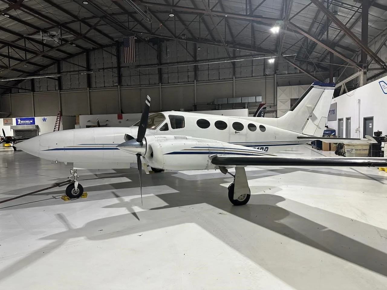 A white Cessna 414 Chancellor twin-engine aircraft with blue stripes is parked inside a hangar.