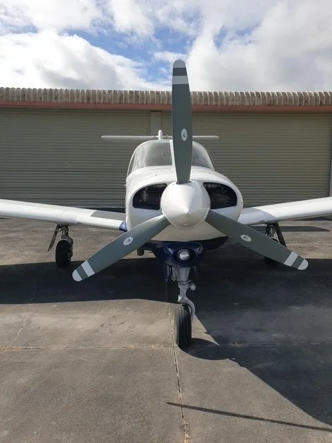 A front view of a white and blue Piper Arrow aircraft with its propeller facing forward, parked on a tarmac.