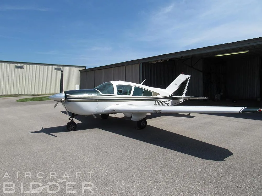 A white and dark green single-engine aircraft N9612E parked on a tarmac in front of hangars under a blue sky.