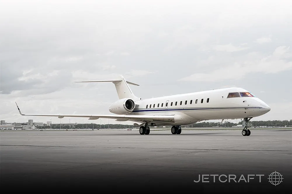 A white Bombardier Challenger 605 business jet with a blue stripe on the tarmac under a cloudy sky.
