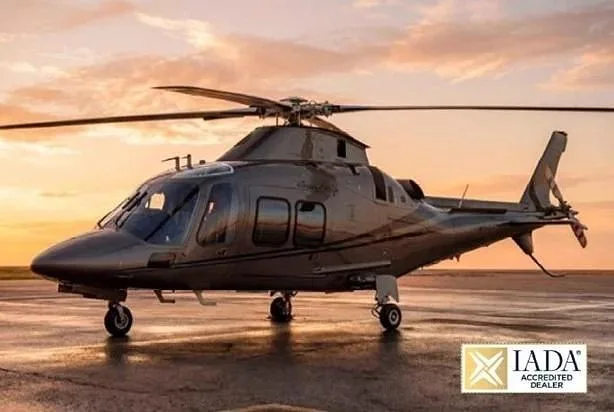A dark grey executive helicopter is parked on a wet tarmac at sunset.