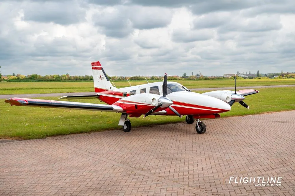A red and white twin-engine Piper Seneca aircraft parked on a paved surface, viewed from the front-right.