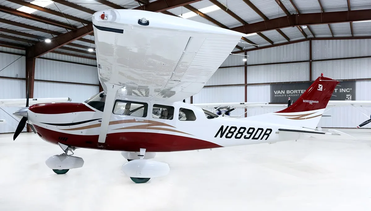 A white Cessna Stationair with red and gold stripes is parked in a hangar.