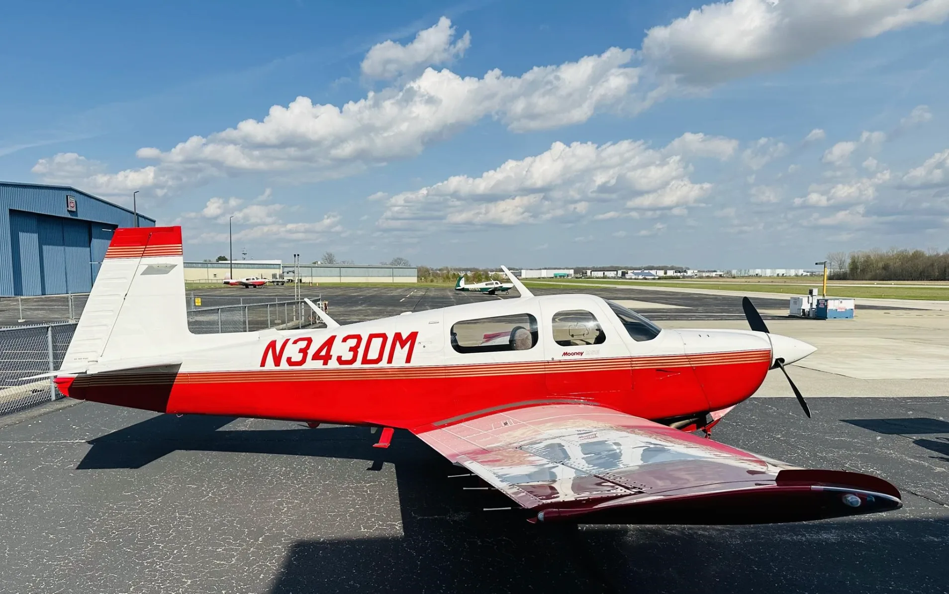 A side view of a red and white Mooney aircraft (N343DM) parked on an airport tarmac under a blue sky.