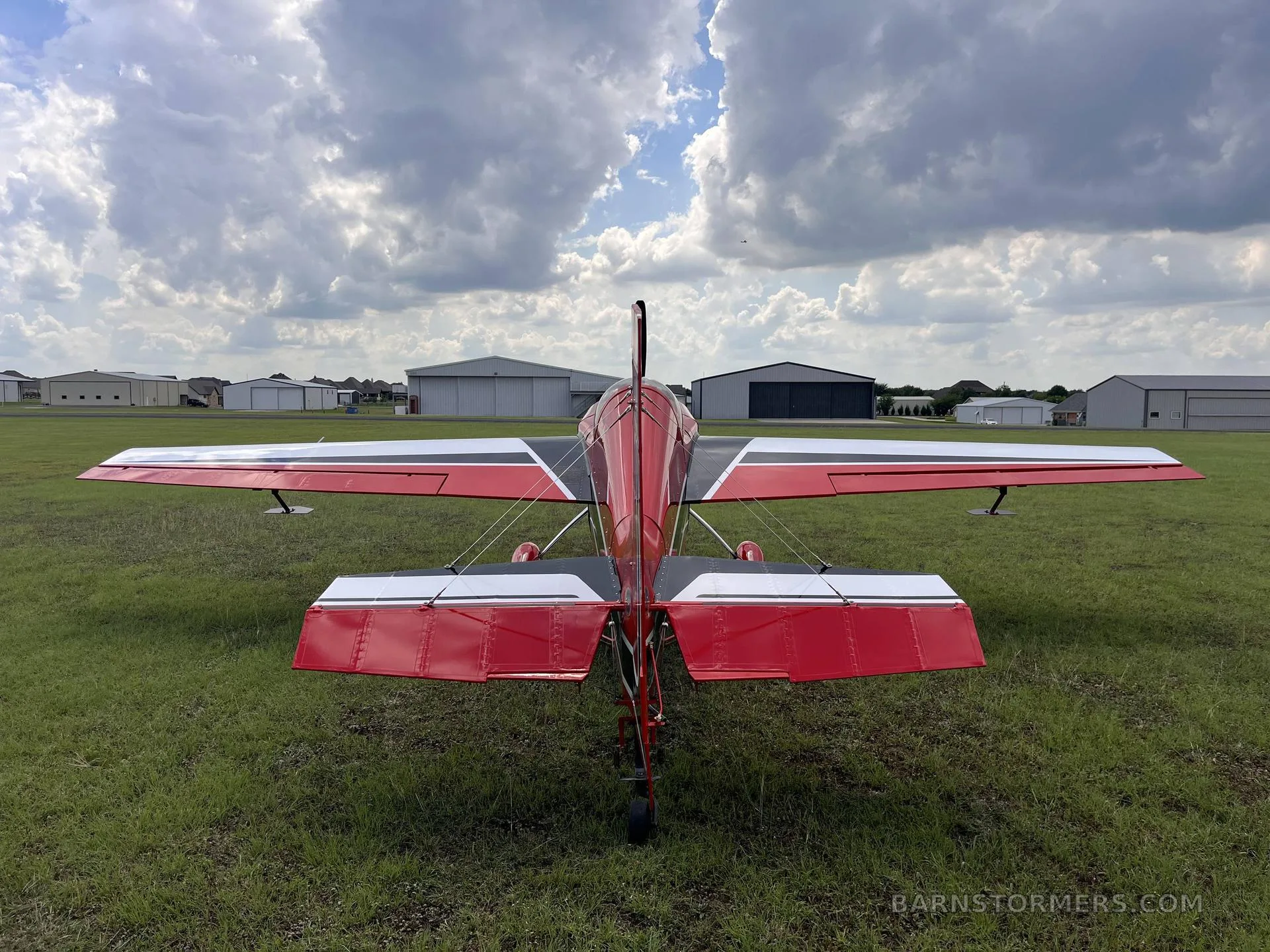 A rear view of a red and white aerobatic aircraft, N300CB, parked on a grassy airfield with hangars in the distance.